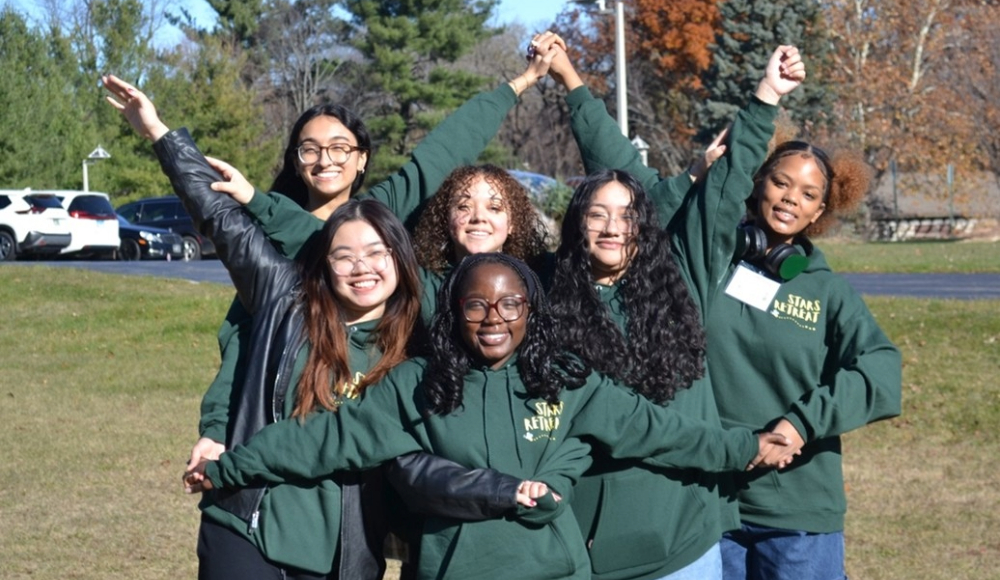 Several young women in green sweatshirts, posing with their arms together in a star formation with cheerful expressions in an outside setting
